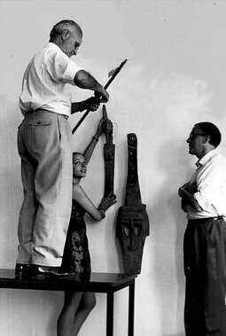 Debout sur la table, le professeur Dominique Zahan travaille à la mise en place d'une exposition d'objets d'art africains avec Erica Haddouf-Nachez (1967)